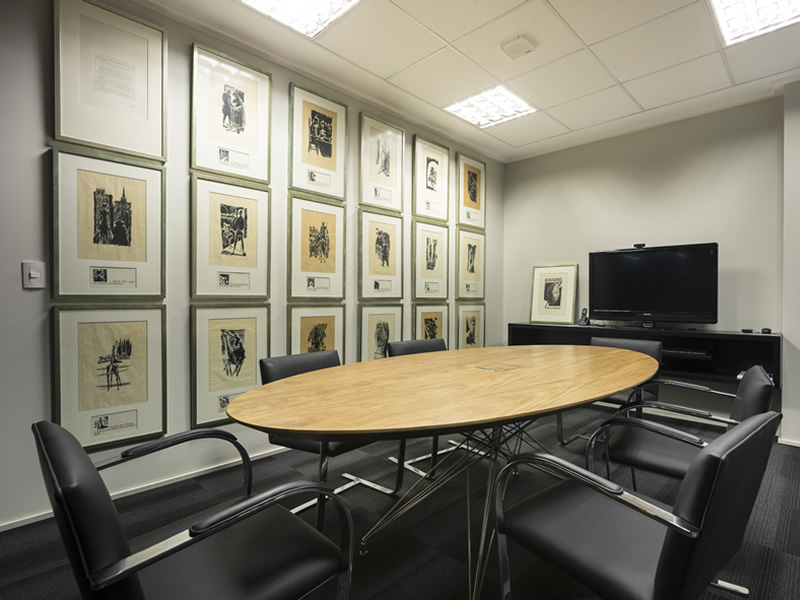 A imagem mostra uma sala de reuniões moderna e sofisticada. A parede principal é adornada por uma galeria de quadros em molduras finas, criand o um ambiente elegante e convidativo. Uma mesa oval de madeira clara, com cadeiras estofadas em couro preto, ocupa o centro da sala, proporcionando um espaço confortável para reuniões e discussões. O piso em carpete cinza claro e o teto com iluminação embu tida completam o design minimalista e contemporâneo do ambiente. A presença de uma televisão na parede oposta à galeria de quadros sugere que a sala também pode ser utilizada para apresentações e videoconferências. No geral, a imagem transmite uma sensação de profissionalismo e modernidade, ideal para um ambiente de negócios.