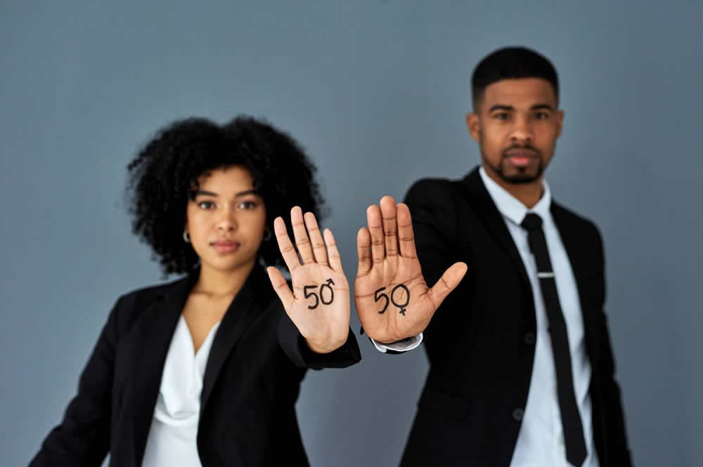 Imagem de um home e uma mulher, ambos negros, vestindo terno preto e camisa branca. Eles estão com as palmas da mão para frente e nelas há um número 